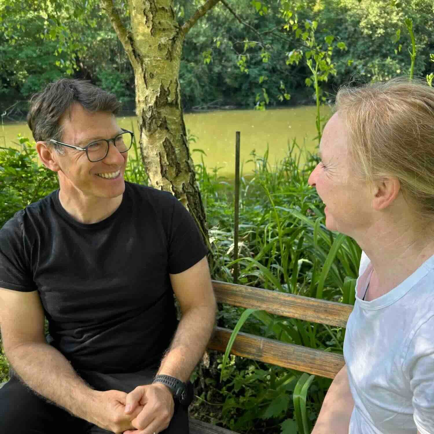 Échange en séance de coaching à Neuchâtel – relation humaine et écoute Coach et cliente en pleine discussion bienveillante sur un banc au bord d’une rivière à Neuchâtel, entourés de verdure