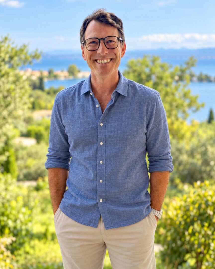 David Juncker souriant, en chemise bleue et pantalon beige — tenue et arrière-plan retouchés par IA.